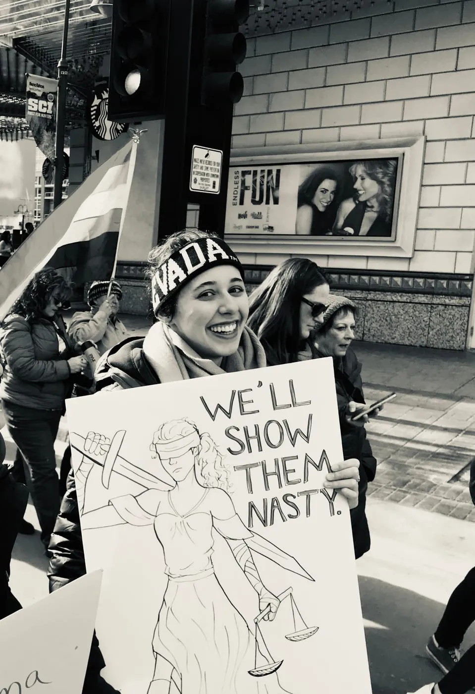 Erica smiling at a march, holding a hand-drawn sign of Lady Justice that reads We'll show them nasty